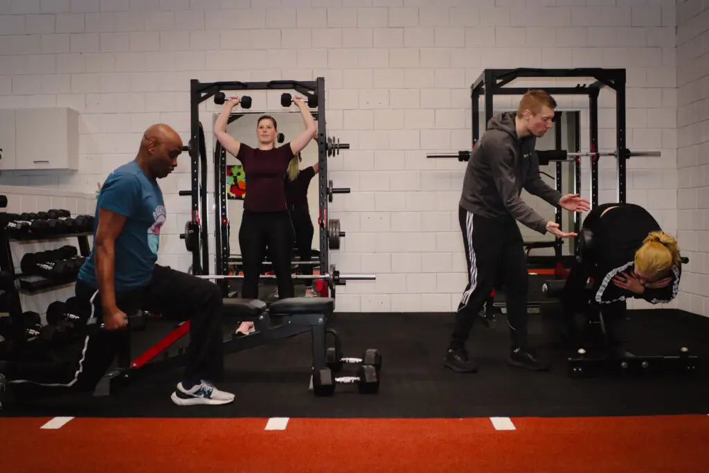 Trainer van BerkSports begeleidt twee sporters bij dumbbell-oefeningen bij het squatrack