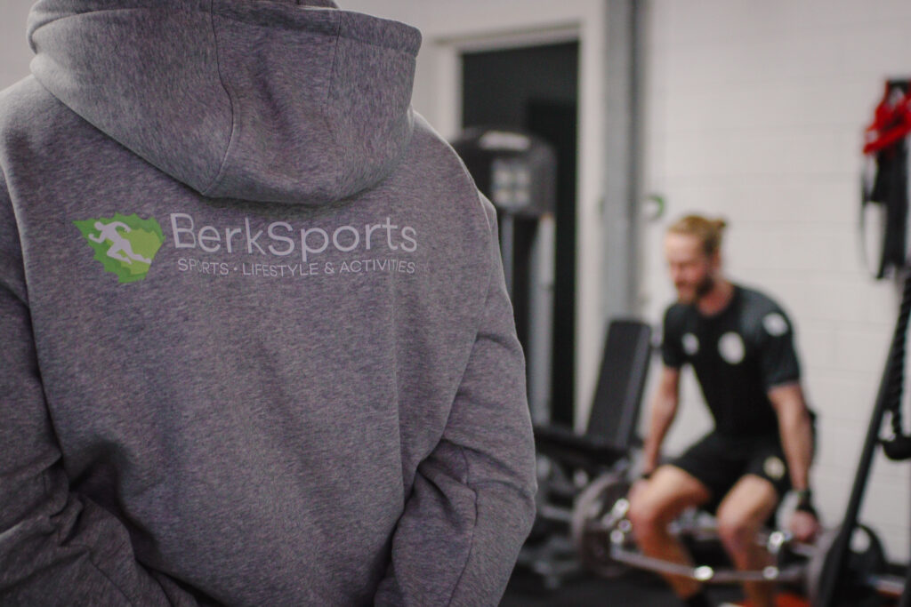 A BerkSports trainer in a hoodie seen from behind; in the background someone is training with a barbell